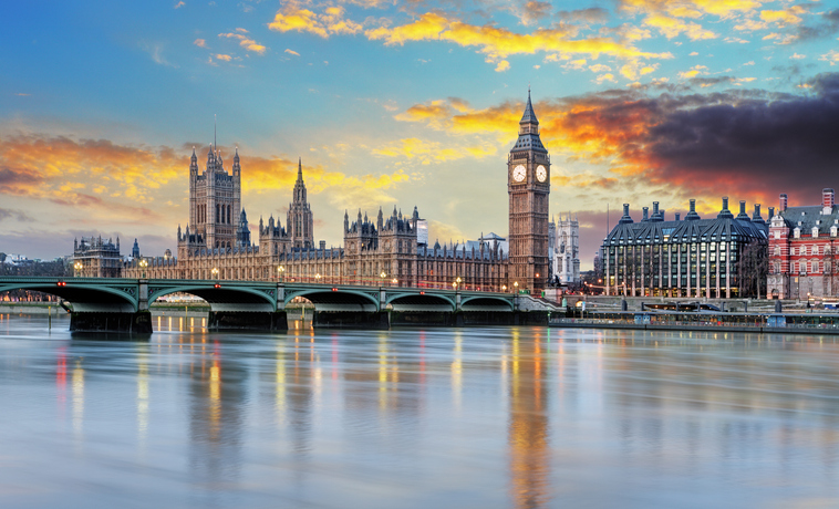 London’s Palace of Westminster and Big Ben beside the River Thames, with Westminster Bridge in the foreground and a colorful sunset sky reflecting on the water.