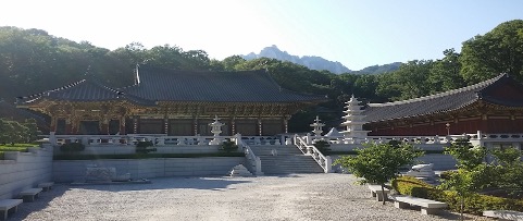 Traditional Korean temple complex with ornate wooden buildings, stone pagodas, and landscaped grounds, backed by forested mountains under a clear blue sky.