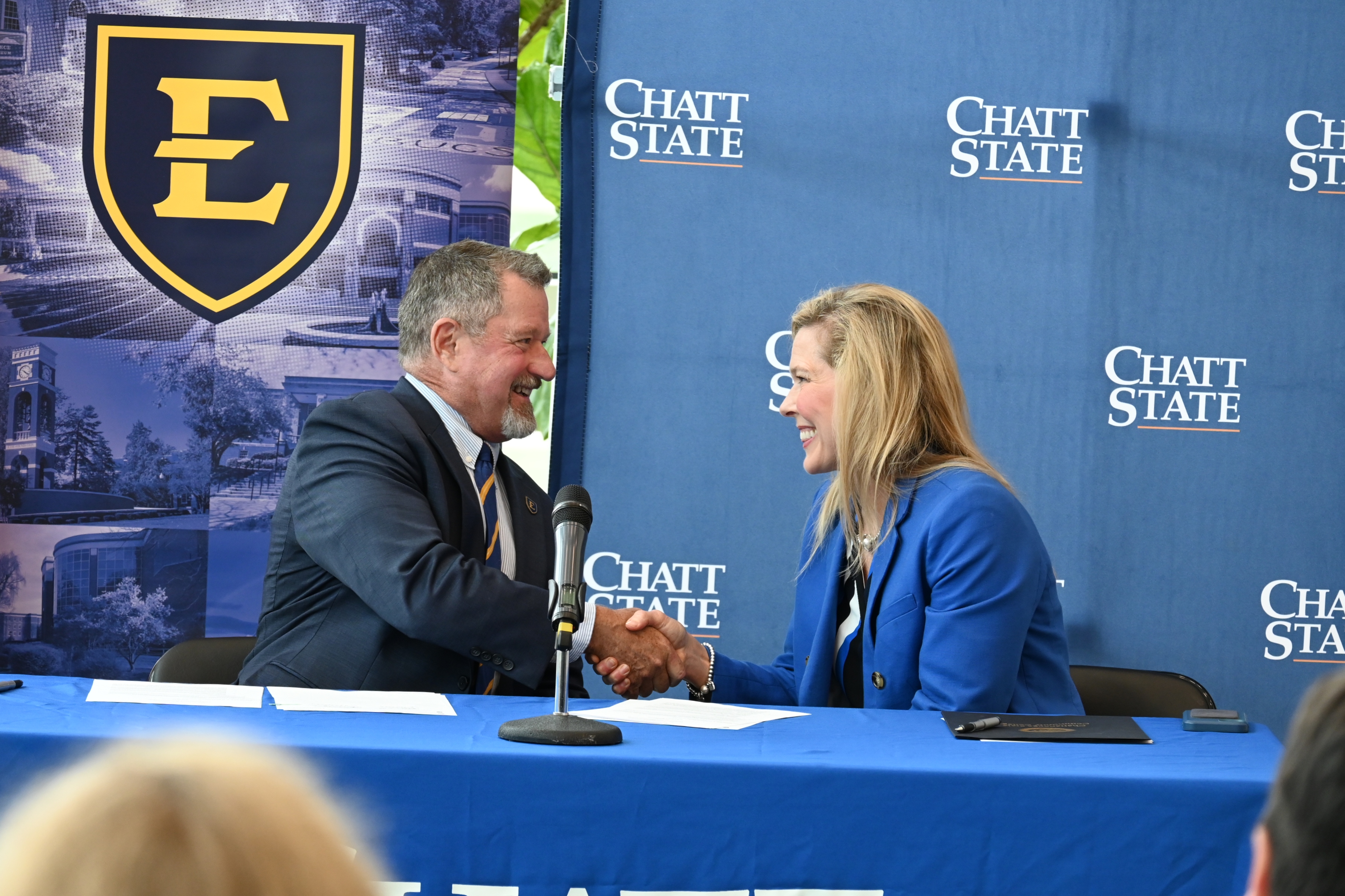 A man in a suit shakes the hand of a woman in a suit in front of ETSU and ChattState backdrops.