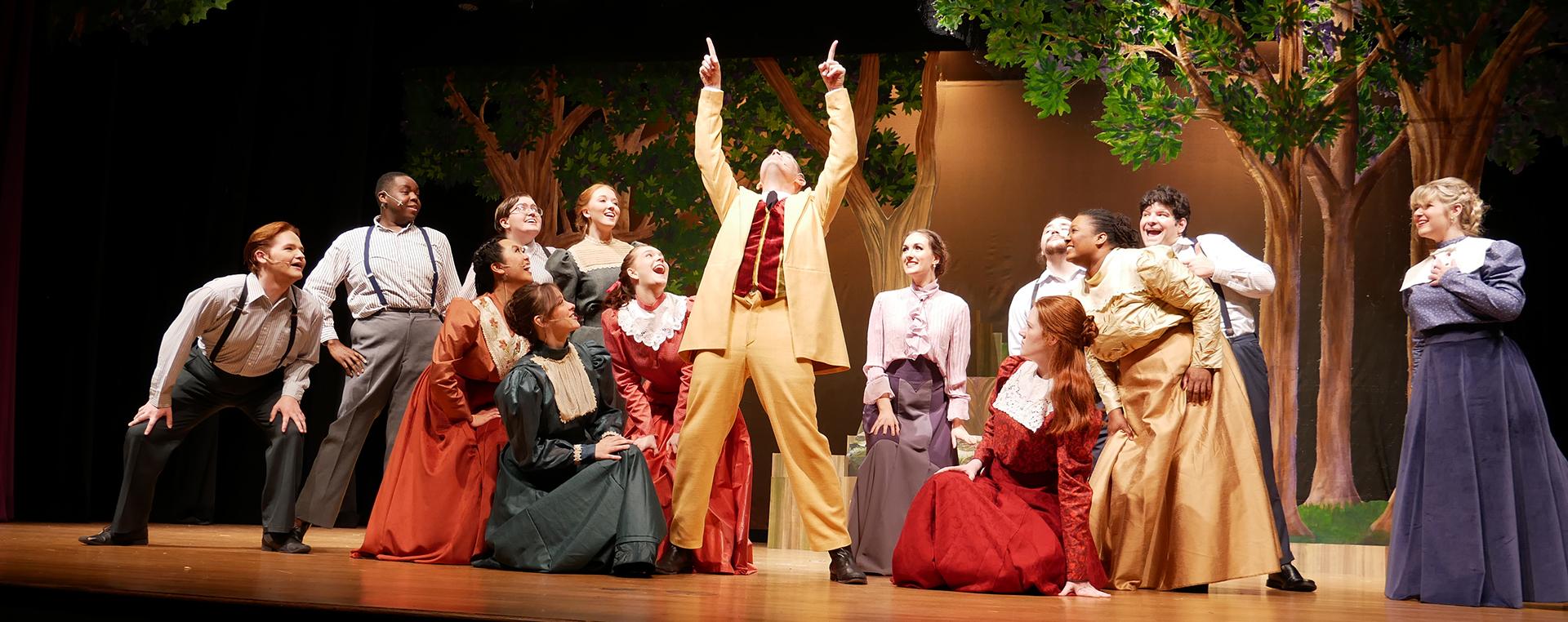 14 actors and actresses on stage all looking at the man in the center