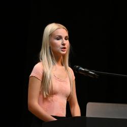 A blonde woman in a pink shirt speaks into a microphone.