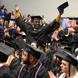 A Chattanooga State graduation in regalia stands with his arms raised surrounded by fellow graduates. A Chattanooga State graduation in regalia stands with his arms raised surrounded by fellow graduates.