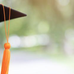 Partial view of an ambiguous student's face wearing a black mortarboard and orange tassle. Out of focus trees are visible in the background.
