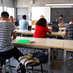 A group of students in a classroom face a teacher and blackboard with electrical diagrams on it. A group of students in a classroom face a teacher and blackboard with electrical diagrams on it.