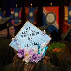 A student attending graduation wears a mortarboard that reads "We Are the Dreamers of Dreams." A student attending graduation wears a mortarboard that reads "We Are the Dreamers of Dreams."