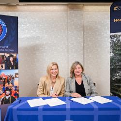 Two blonde women in business casual clothing sign articulation agreements on a blue table flanked by signs promoting ChattState and UTC.