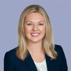 Tiffanie Robinson, a blonde woman in a black coat, smiles in front of a light blue background.