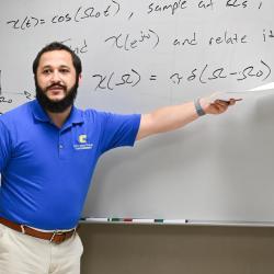A man in a blue polo shirt points to a white board.