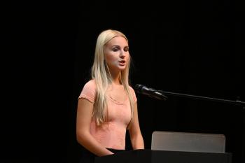 A blonde woman in a pink shirt speaks into a microphone.