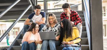 Stock photo of five college-aged students sitting on stairs looking at a laptop.