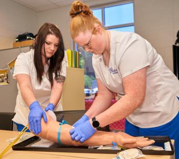 Two female nursing students in white scrubs practicing inserting an IV needle into a plastic prosthetic arm. Two female nursing students in white scrubs practicing inserting an IV needle into a plastic prosthetic arm.