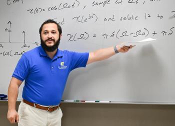 A man in a blue polo shirt points to a white board.