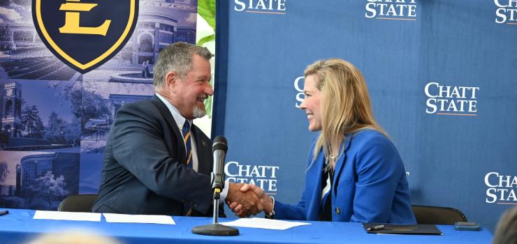 A man in a suit shakes the hand of a woman in a suit in front of ETSU and ChattState backdrops.