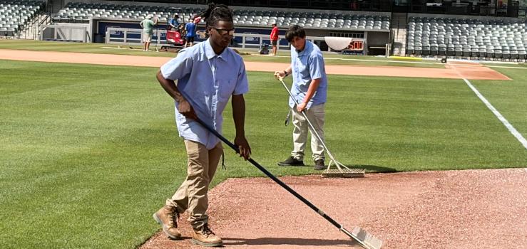 Two male students in workware use rakes to even out gravel on a baseball field.